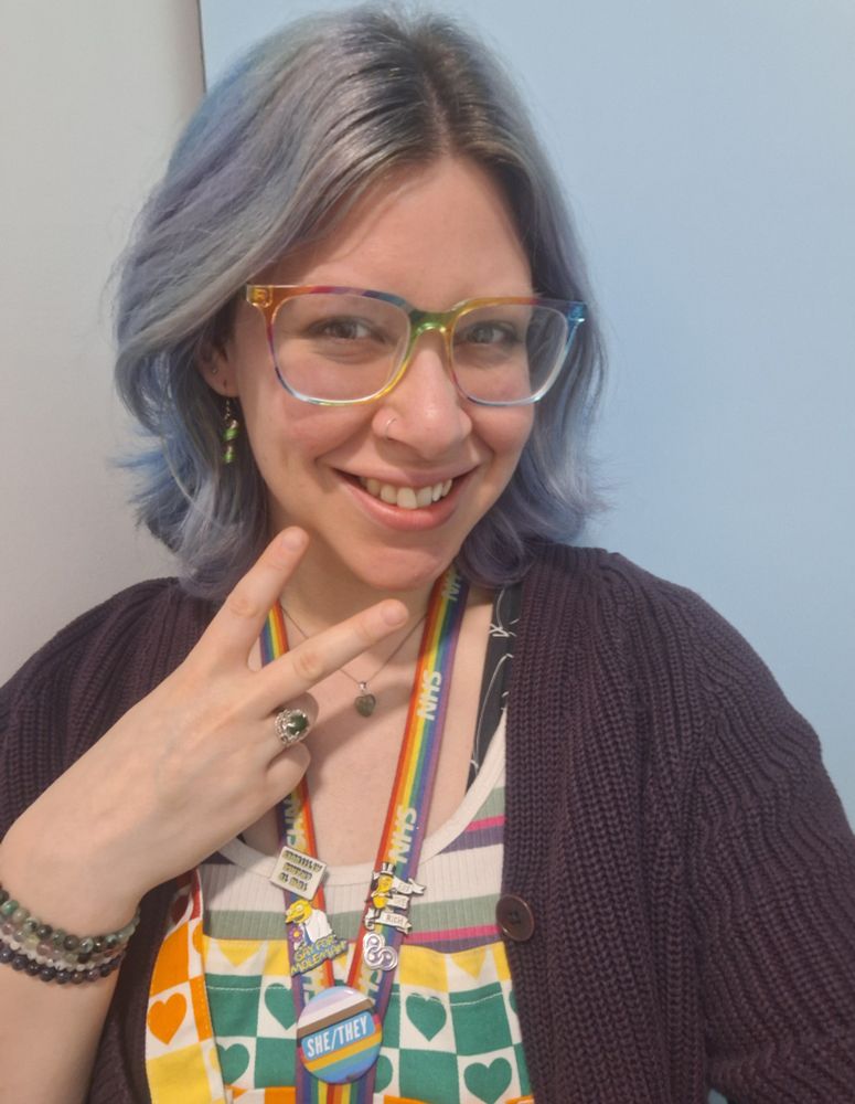 A selfie photo of Lady G, a white genderqueer person in their mid thirties. They have short purple hair, rainbow glasses and a she/They pronoun badge. They are wearing rainbow dungarees and purple white and green jewellery. 
