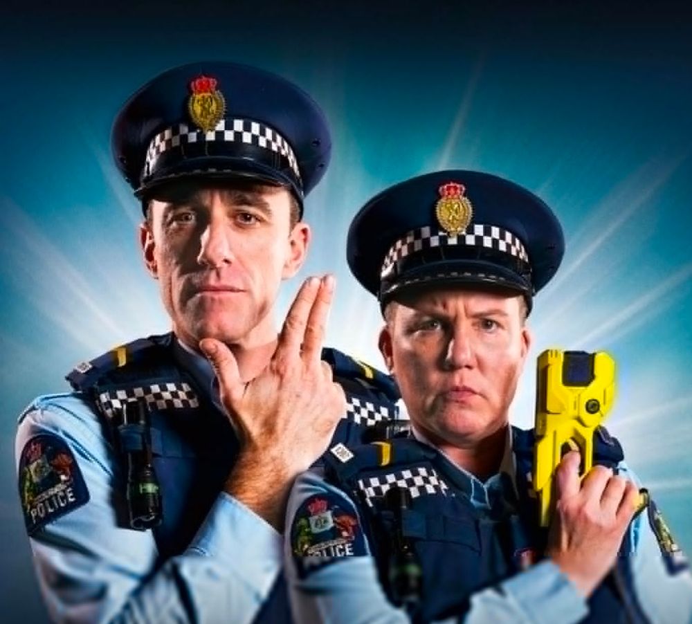 Officers Minogue and O'Leary from Wellington Paranormal are looking very seriously straight at the camera. Minogue is holding up his fingers like a gun and O'Leary is holding up a tazer.