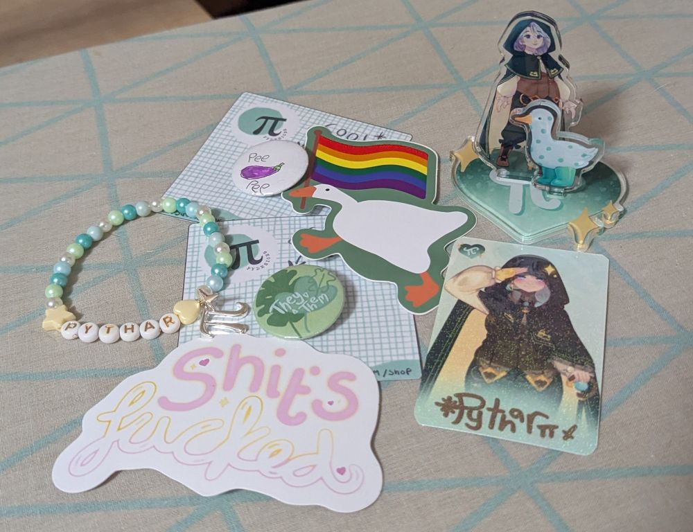 A photo of items from the Py Designs shop, including:
a beaded "pythar" bracelet, a "shit's fucked" sticker, a they/them pronoun badge with a leaf design, a pee pee eggplant badge, a sticker of a goose holding a rainbow flag, a pythar photocard, and a pythar acrylic standee