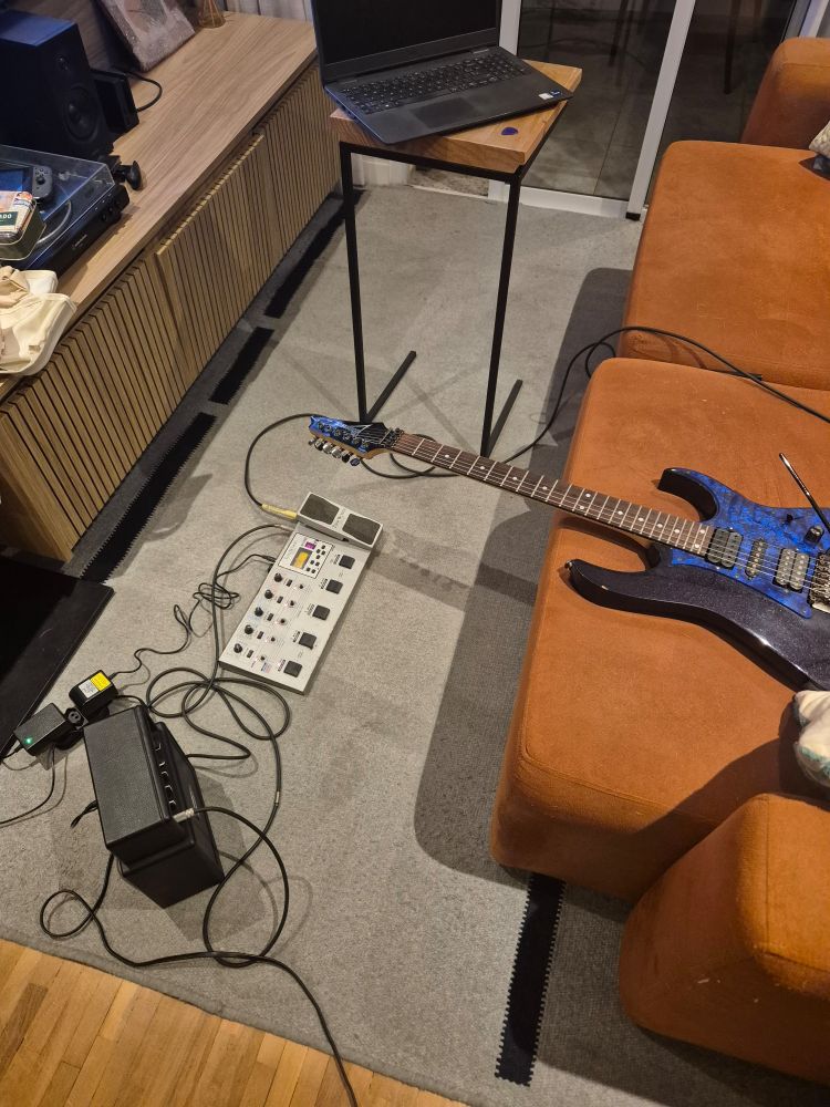 Mini amplificador, uma peladeira, uma guitarra roxa e um notebook numa sala de estar