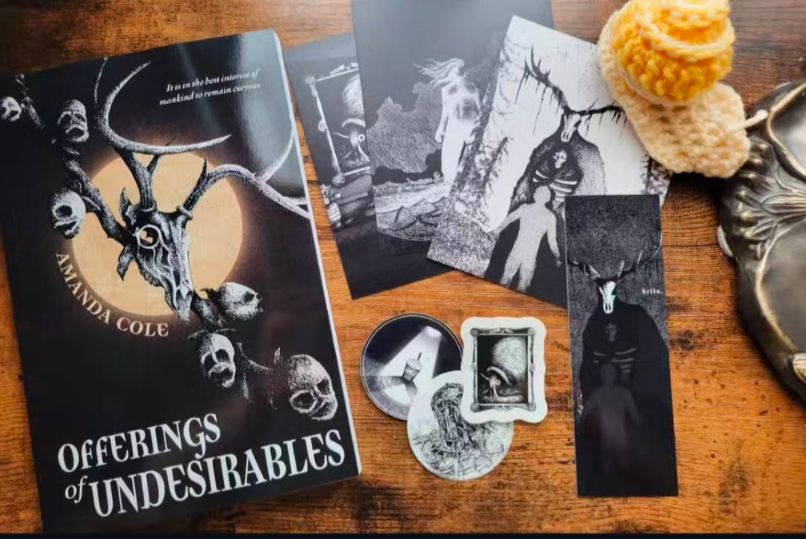 Photo of a book, bookmark, mini art prints and stickers for a giveaway. Book is 'offerings of undesirables' by Amanda Cole