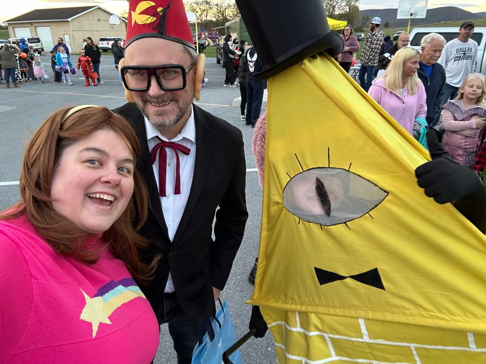 A 40-something dressed as Mabel from Gravity Falls (with a pink sweater with rainbow shooting star, brown hair, and headband), a 40-something dressed as Gruncle Stan (with oversized ears, glasses, a fez, and bow tie), and a child dressed as Bill Cipher (a large yellow 2 dimensional triangle with a large eye, bow tie, bricks, top hat, and cane)