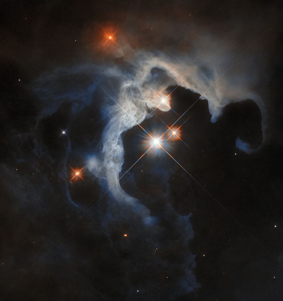 A long, smoky, greyish-blue cloud in the centre of the image curves in an arc around three bright stars, each with long cross-shaped diffraction spikes. The cloud is lit more brightly on the inner side facing the stars, and fades into the dark background on the outer side. A few other stars and points of light surround the cloud: one small star below it has a dark band crossing its centre.