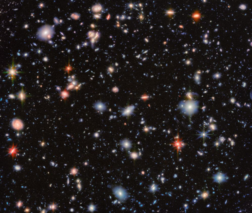 This image from the NASA/ESA/CSA James Webb Space Telescope revisits one of the most iconic regions of the sky, the Hubble Ultra Deep Field. It shows an area of deep space with thousands of galaxies in various shapes and sizes on a black background. Most are circles or ovals, with a few spirals. More distant galaxies are smaller, down to being mere dots, while closer galaxies are larger and some appear to be glowing. Red and orange galaxies contain more dust or more stellar activity.