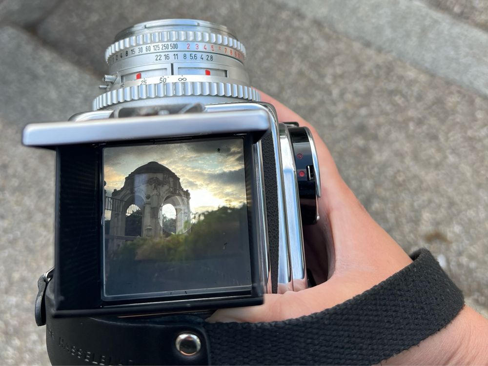 Photograph of a hand holding a camera with a ground glass viewfinder. In the viewfinder is a picture of a dome in the Vienna Stadtpark.