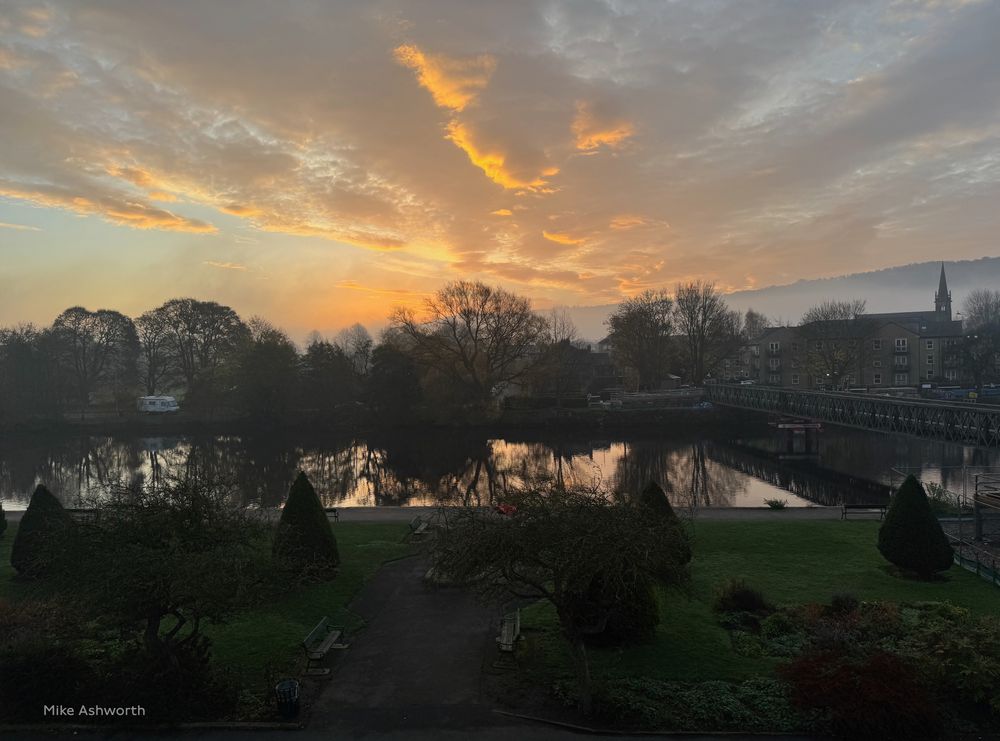 Autumnal view - sun rising through low clouds and mist looking across park land and the River Wharfe in Otley, West Yorkshire. The now bare trees and light are reflected in the water.