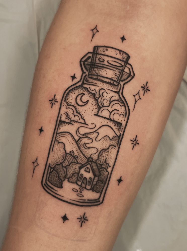 This is a photograph of a fresh tattoo on a person’s forearm. The tattoo depicts a bottle with a cork stopper. Inside the bottle is a landscape scene with a house, trees, bushes, mountains, a moon, and clouds. The tattoo is done in black line work and dot work style. Done by Molly Vee in Seattle, WA. 