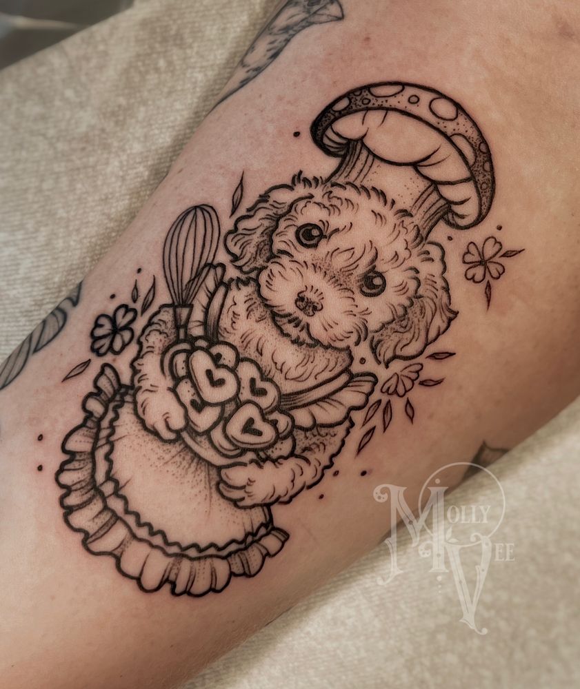 This is a photo of a tattoo on a woman’s arm. The tattoo is done in black line work style and it depicts a labradoodle dog wearing an apron and chefs hat, holding a tray of heart shaped cookies. The tattoo is done by Molly Vee in Seattle WA