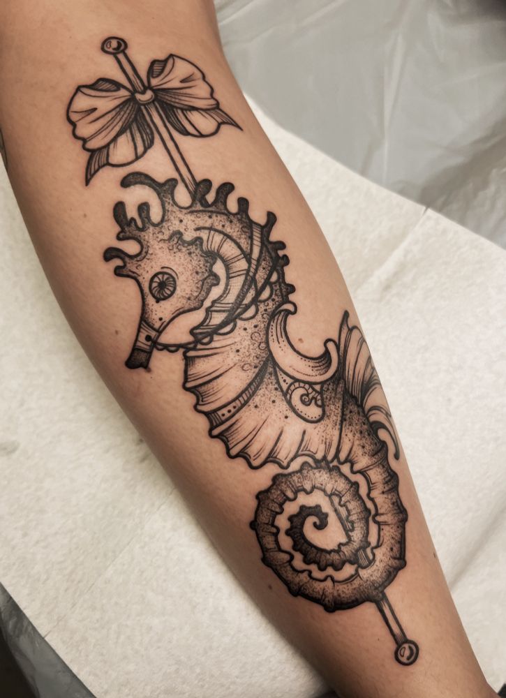 This is a photo of a tattoo on a woman’s leg. The tattoo is done in black line work style with dot work and stippling details. The image is a seahorse done in the style of a carousel horse with a pole and saddle. Done by Molly Vee in Seattle WA.