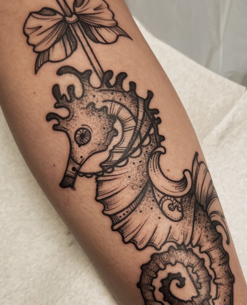 This is a photo of a tattoo on a woman’s leg. The tattoo is done in black line work style with dot work and stippling details. The image is a seahorse done in the style of a carousel horse with a pole and saddle. This image is slightly zoomed in compared to the other image in this post. Done by Molly Vee in Seattle WA.