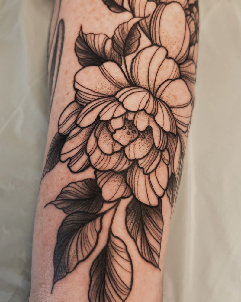 Pictured is a fairly close-up photo of a fluffy rose tattoo on a woman’s forearm. The tattoo is done in black and grey line work with soft black shading and stippling. The leaves are shaded darker than the petals, and there is fine line detail throughout. 