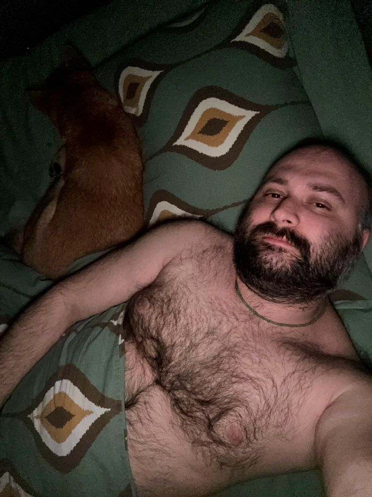 A shirtless me lying in bed next to my shiba pup, both partially covered by a green patterned blanket.
