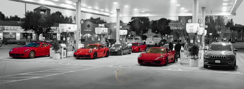 A nighttime gas station scene featuring a striking lineup of luxury sports cars, predominantly Ferraris, with selective color editing that highlights the red vehicles while the rest of the image remains desaturated. The well-lit station has multiple fuel pumps and a few people standing near the cars.

From left to right, the vehicles include:

Ferrari 599 GTB Fiorano – A red grand tourer with a long hood, gold wheels, and a sleek profile. This V12-powered front-engine Ferrari is designed for both speed and comfort.
Porsche 718 Cayman – A mid-engine sports coupe with black wheels and an aggressive stance, known for its precise handling and balanced performance.
Ferrari 458 Italia – A mid-engine supercar with aerodynamic curves and sharp lines, one of the last naturally aspirated V8 Ferraris.
Ferrari 488 GTB or F8 Tributo – Another red Ferrari positioned further back, featuring a twin-turbo V8 engine and a design inspired by Formula 1 technology.
Ferrari F430 – A prominent red Ferrari in the foreground with smooth curves and an iconic front grille, a mid-2000s supercar that introduced electronic differential technology.
Jeep Cherokee – A silver SUV parked at the rightmost fuel pump, providing a stark contrast to the high-performance sports cars with its practical and rugged design.
The gas station’s background includes a UPS Store and a coffee shop, with trees and buildings visible under the night sky. Bright overhead lights illuminate the cars, reflecting off their polished exteriors. The image’s selective coloring draws attention to the vibrant red supercars, emphasizing their sleek and exotic nature against the monochrome surroundings.