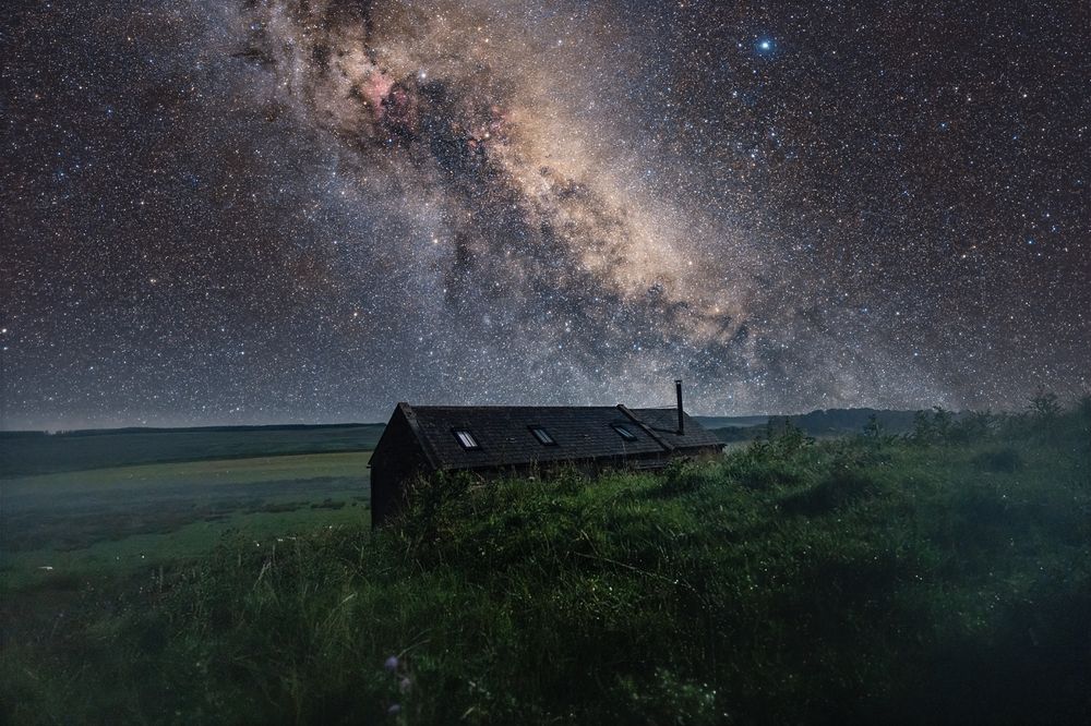 A wide-field night photograph of the Milky Way arching across the sky, with the Cygnus star cloud and its dark dust lanes clearly visible. A small dark-roofed cottage sits in the foreground on a grassy hillside, while the bright star Deneb shines above the galactic band. The image reveals dense star fields, reddish nebula patches, and intricate dark structures cutting through the glowing galactic plane.

