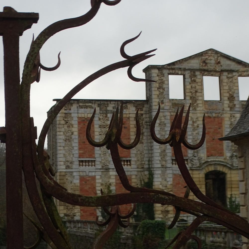 The façade of the Château of Thury-Harcourt, with two floors of bricked up windows and a top floor of three empty window frames, viewed through the ironwork of the Château gates.