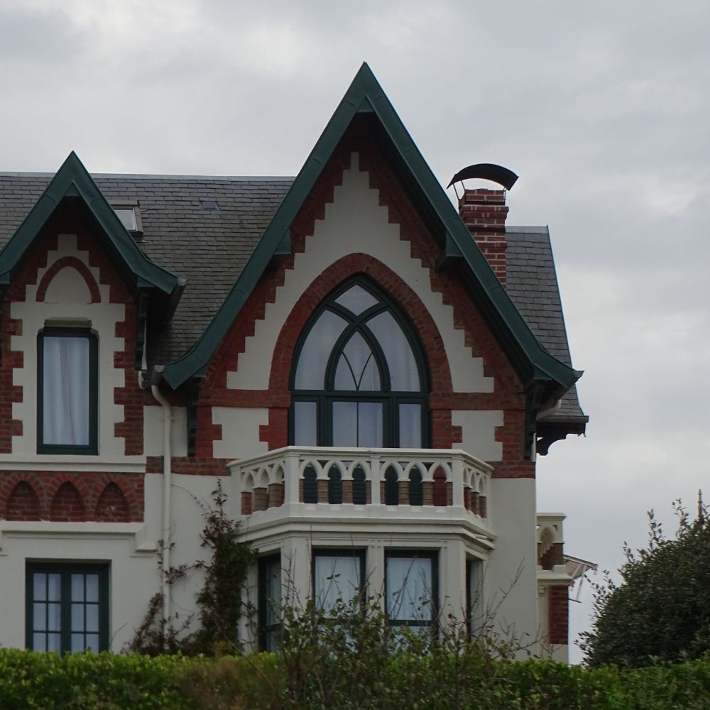A cropped image of a decorative brick house with a large arc-shaped window on the first floor which leads onto a balcony where the arc shape is repeated in the stone balustrade