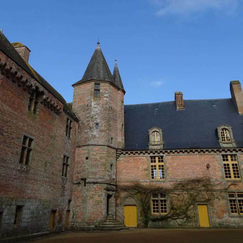 The interior courtyard of a red-brick castle. There is a tall tower with a conical slate roof with wings of the castle on either side of it. The window frames and doors on the three floors are painted mustard yellow and the sky is blue.