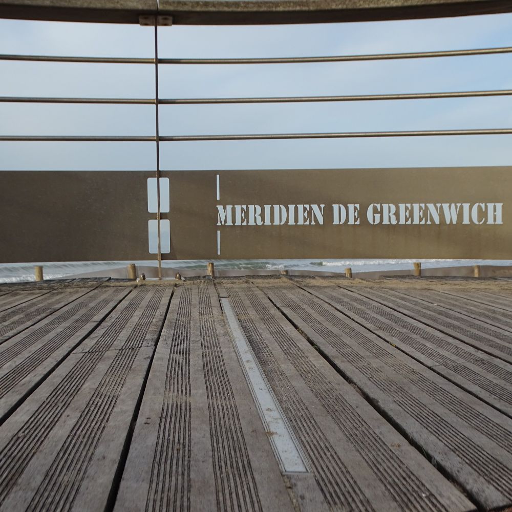 View across some decking to metal railings. On the railings is a sign saying "Meridien de Greenwich" and a marker to show where the meridian passes.
