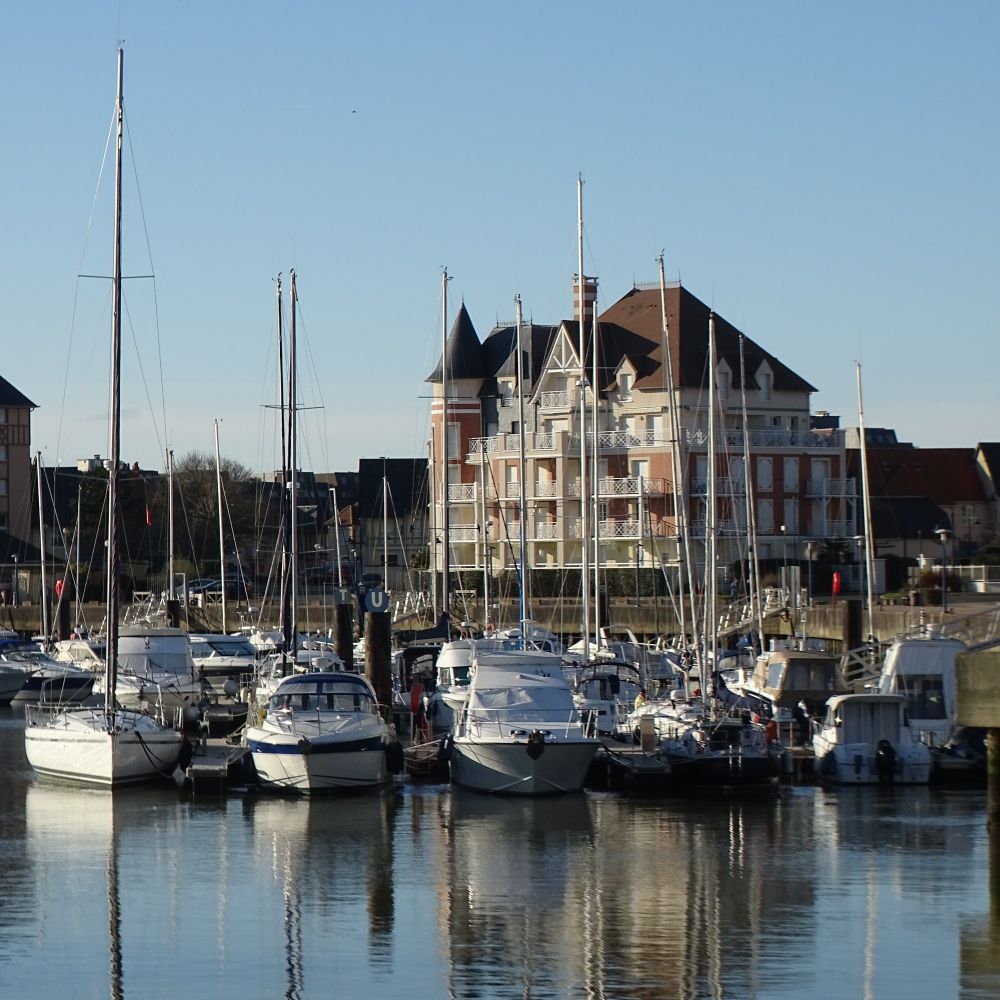 Sailing boats in a modern marina with a large residential building in a Normandy style in the background, with turrets and lots of eaves, under a clear blue sky.