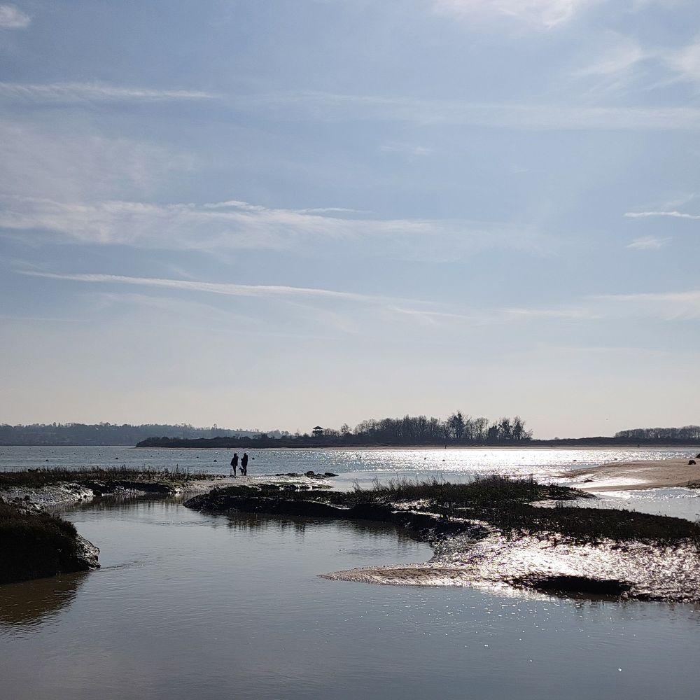Wet sand and mud in a tidal estuary reflect the sunlight. Two distant silhouetted figures walk along a sandbank. The sky has wispy clouds in it.