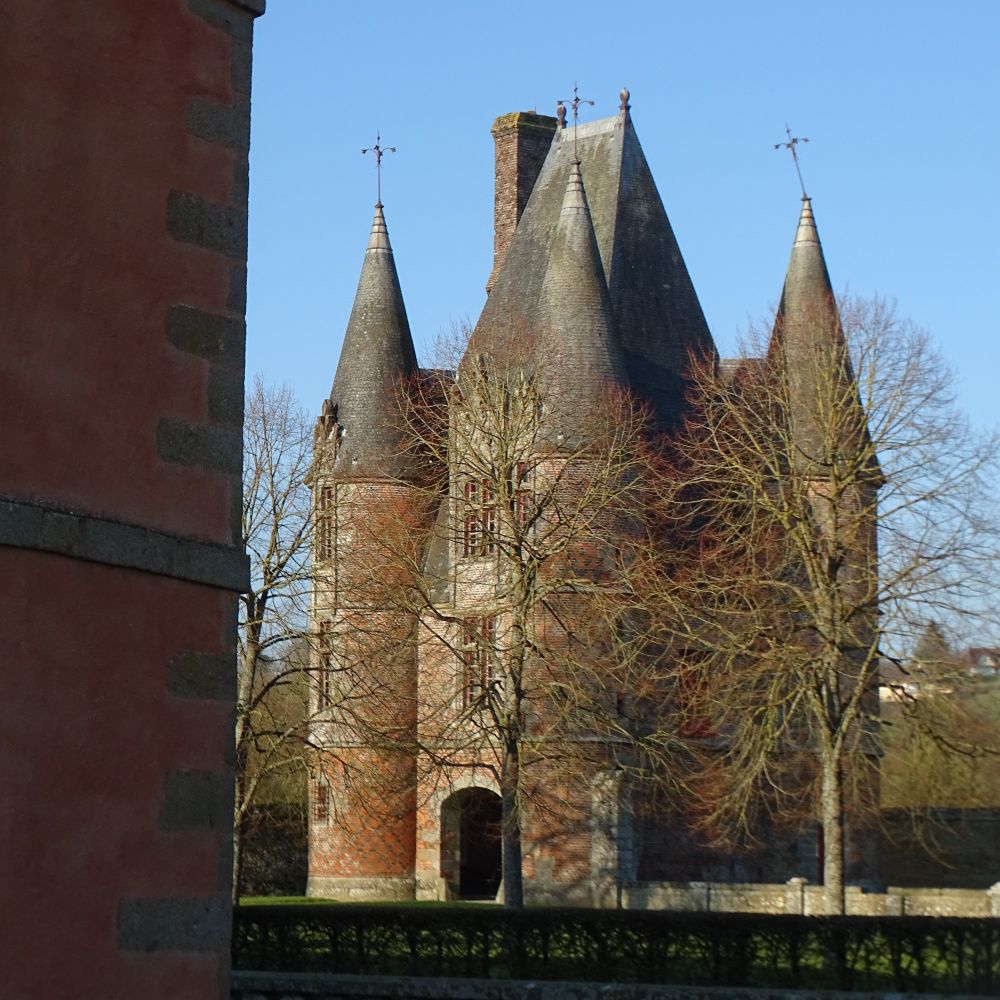 A view of the red-brick gatehouse taken from behind a corner of another building and through some trees with no leaves. The gatehouse has three floors and round towers on each corner with conical slate roofs. The central section has a steep slate roof. One of the weather vanes at the top of the turrets is not straight.