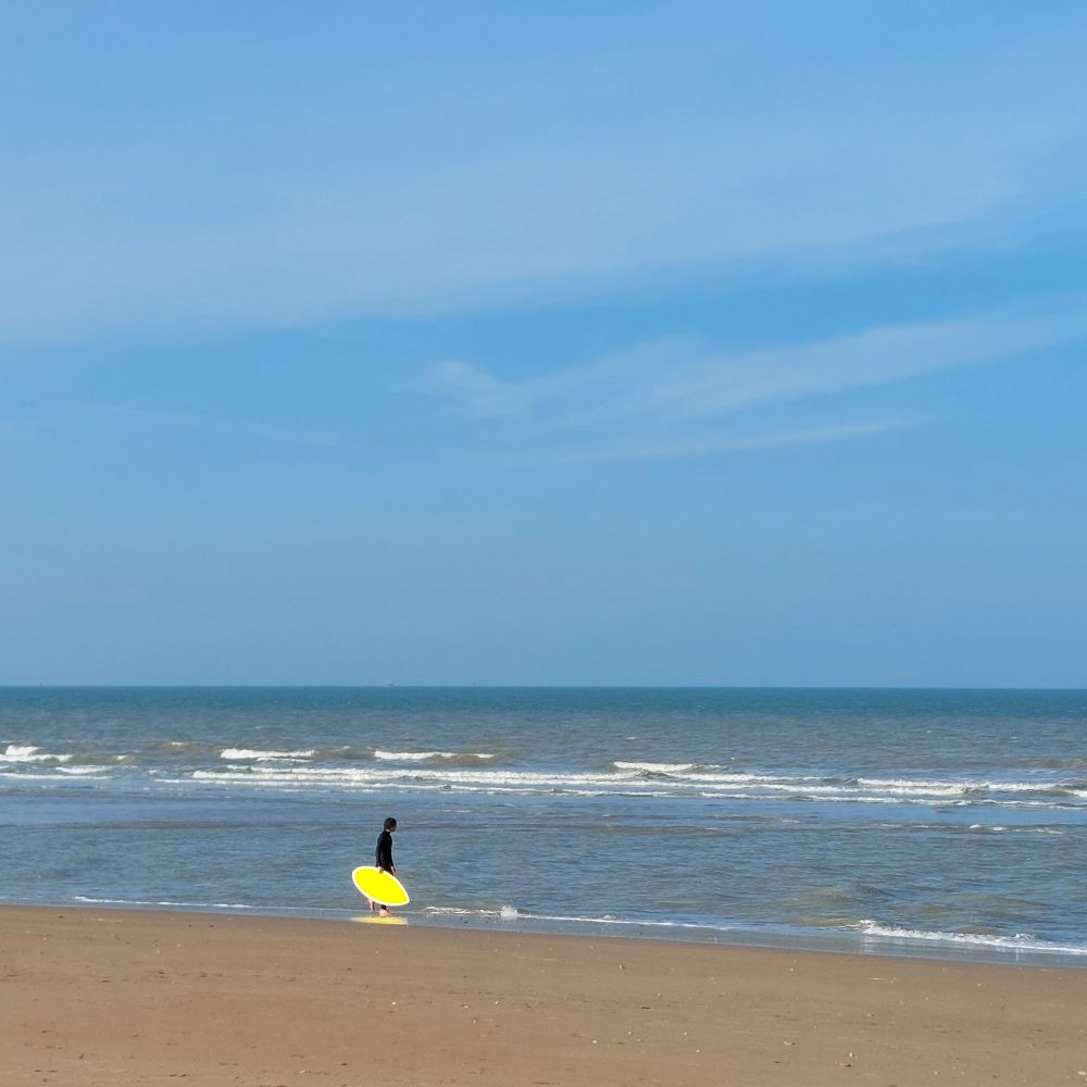 A sandy beach with some small breaking waves under a blue sky. In the middle ground is a figure in a black wetsuit at the edge of the sea, carrying a small, lemon-yellow surfboard.