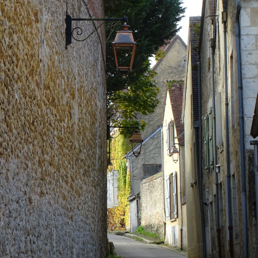 A narrow street with a tall stone wall on the left and stone houses on the right. Old-fashioned street lamps are attached to the wall and there is sunlight at the end of the street