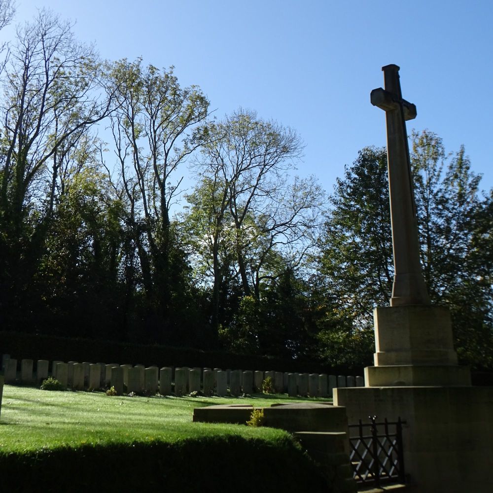A small military cemetery in partial sunlight and surrounded by trees with a tall stone cross and rows of white tombstones.