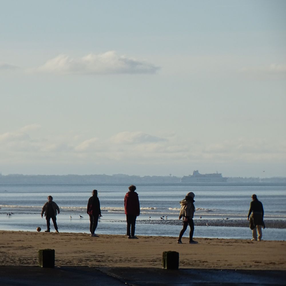 A sandy beach with five people silhouetted against the sea. The sky is clear but hazy, and a ferry is visible against the horizon in the distance. 
