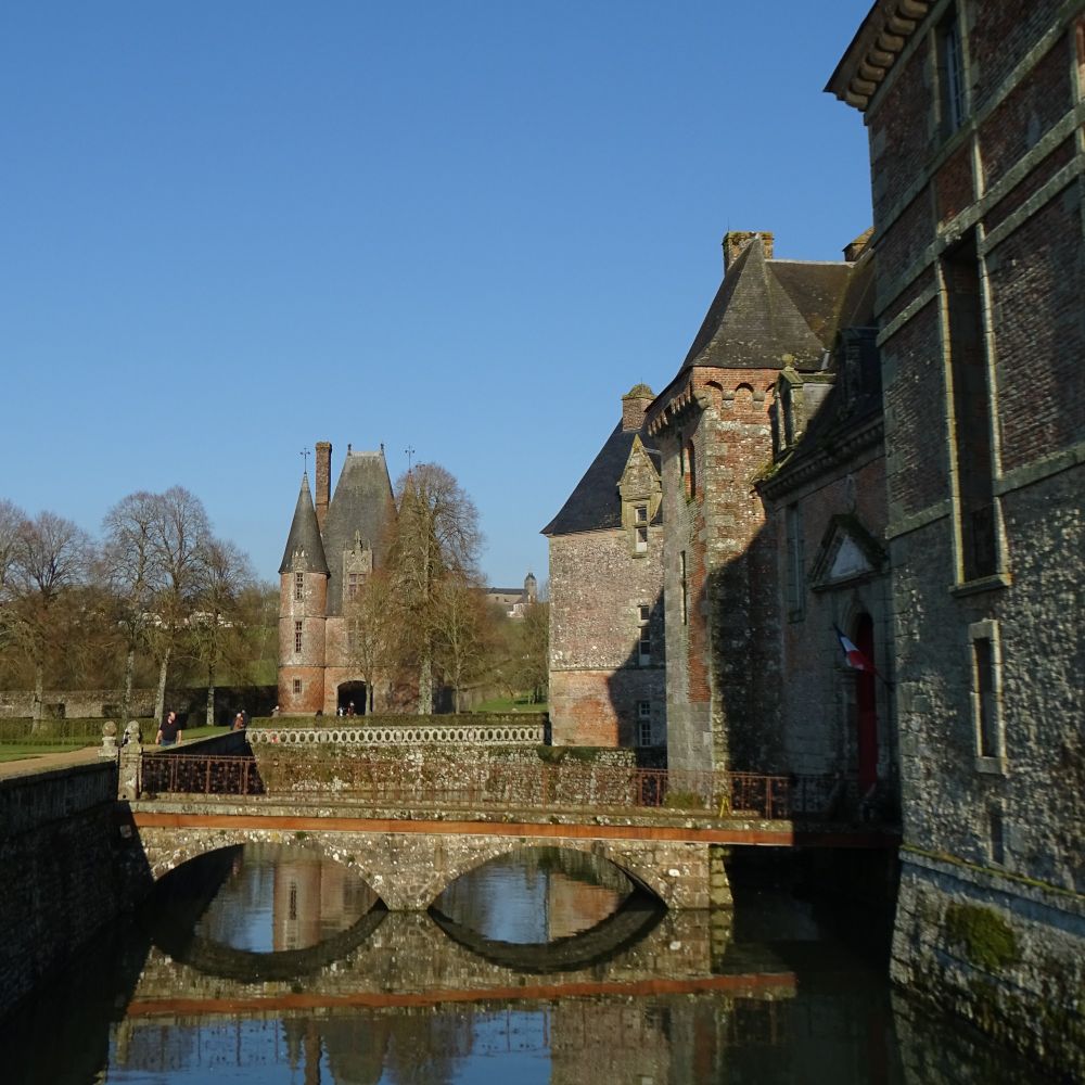 A fortified red-brick castle is on the right of the photo and is surrounded by a moat. A bridge with two arches crosses the moat from left to right. In the distance on the left of the image is the red-brick gatehouse and the sky is blue.