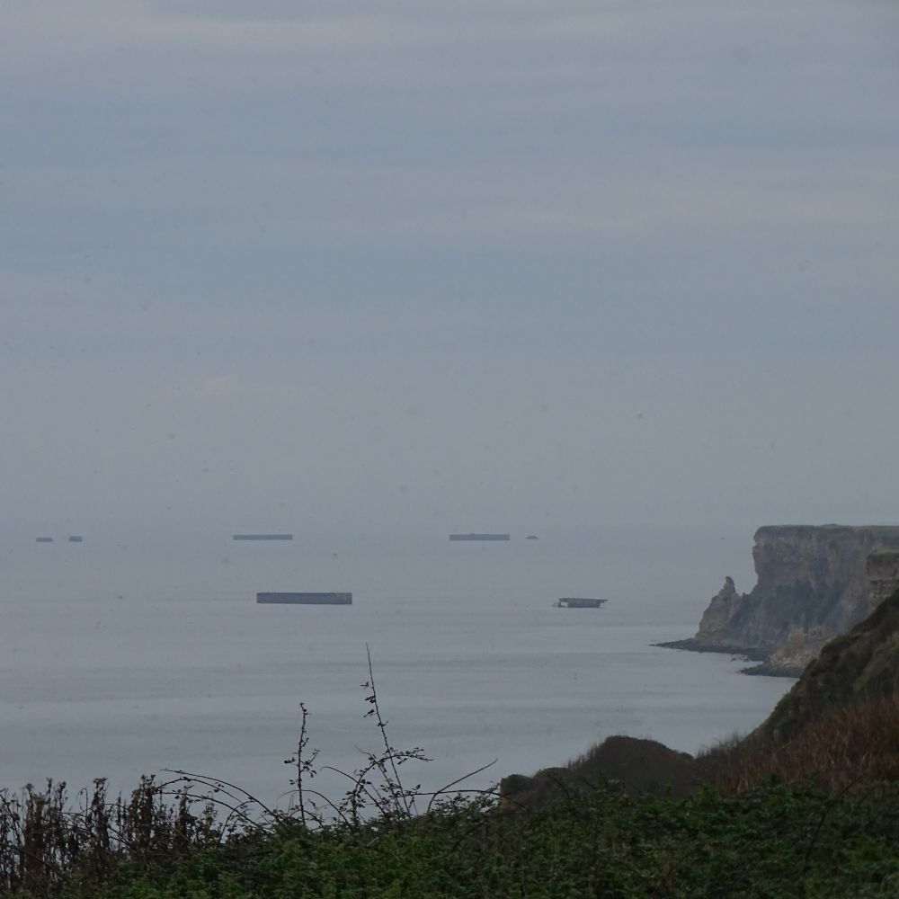View of a coastline with cliffs in the distance and remains of the artificial port at Arromanches in the sea