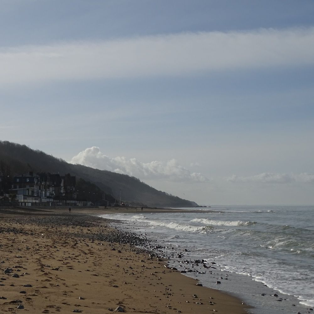 A sandy beach scattered with stones and with waves breaking from the right. The coastline leads into the distance, under a hazy blue sky
