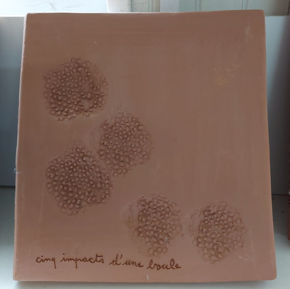 Œuvre en terre cuite rouge d'à peu près 20 x 20 cm avec cinq empreintes rondes et un texte manuscrit qui dit "cinq impacts d'une boule"