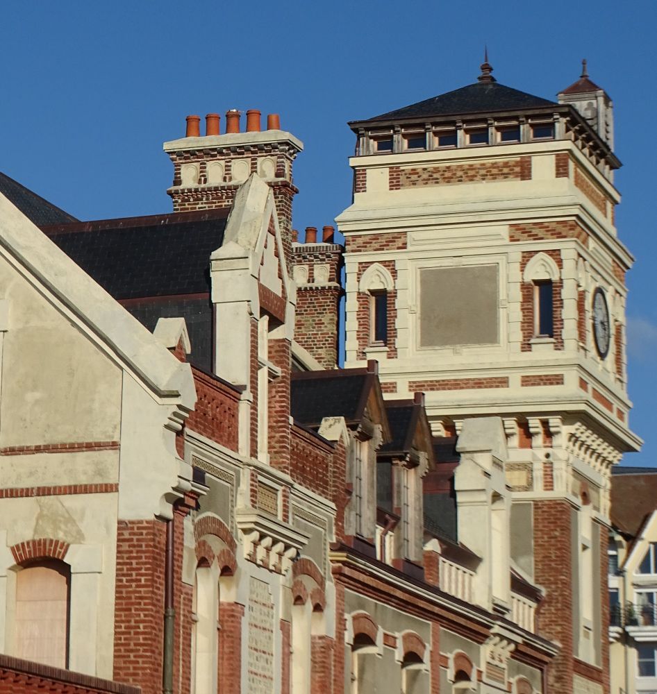 An industrial building from the 1890s with red brick and white stone, with lots of windows and chimneys and a tall square clock tower, all against a clear blue sky.