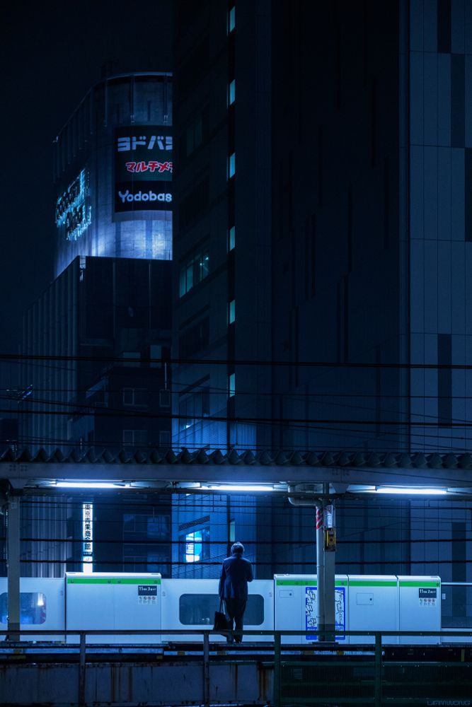 A businessman (salaryman) stands on an empty platform waiting for the last train home, in Tokyo’s Electric Town: Akihabara, Japan. The man is stood in the middle of the frame, captured from behind. The image is vertical with a cold blue sterile feeling to it. Above him and in the distance are large modern buildings, one sign can be seen briefly - YODOBASHI - one of the worlds largest electronic stores. The lights are off. He holds a briefcase and is lit up by halogen lights overhead. Overhead wires and buildings tower over him, he makes up a tenth of the image in scale. At first glance it may look like an illustration due to the colour treatment.