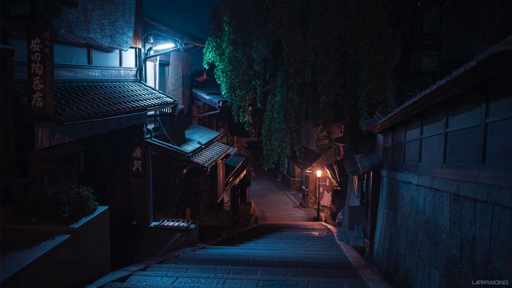 Photographs by Liam Wong of Kyoto at night. An empty scene with traditional Japanese buildings lit up with soft street lights in warm orange colors. The image is mostly blue in color. A large overhanging tree can be seen in the middle of the frame.