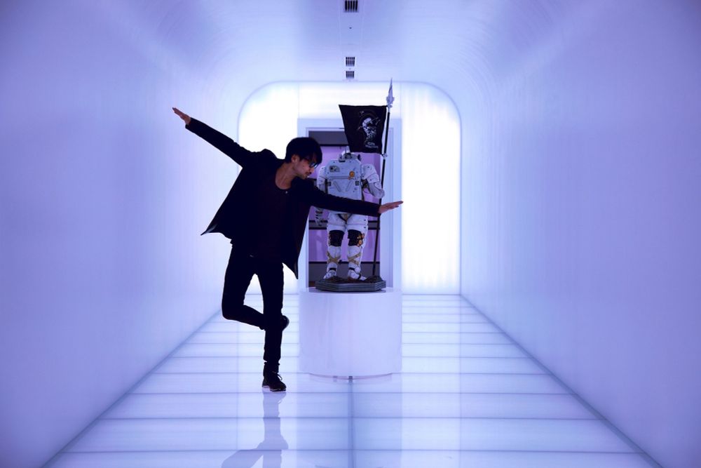 Hideo Kojima in a bright sci-fi styled hallway mid-dancing pose with his company mascot, Ludens behind him. 