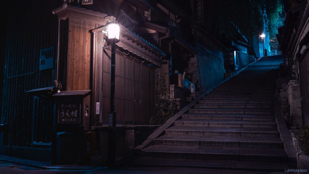 Photographs by Liam Wong of Kyoto at night. A large stairway leading upwards next to an old wooden traditional Japanese building. The image is moody to emphasise the emptiness with brown and blue shades. Towards the top right of the image, the stairway ends and is lit up, with trees overhanging.