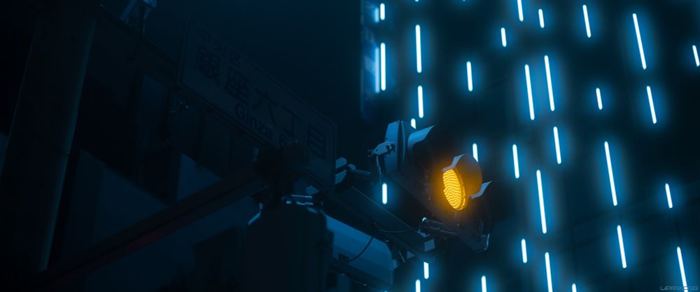 A traffic light and highway sign which reads 'GINZA'. The light is yellow and is glowing. In the background are the rain-like neon light strips which are hazy with a light blue tint.