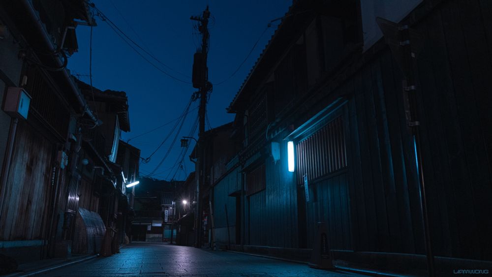 Photographs by Liam Wong of Kyoto in the early hours. Old wooden Japanese traditional buildings. The camera is capturing the scene from ground level, looking up - the blue sky is cut up by the silhouette of an overhead powerline. The image is a cold blue tone.