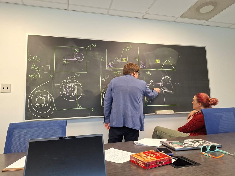 A man draws and explains a series of complicated models on a blackboard while a woman watches.