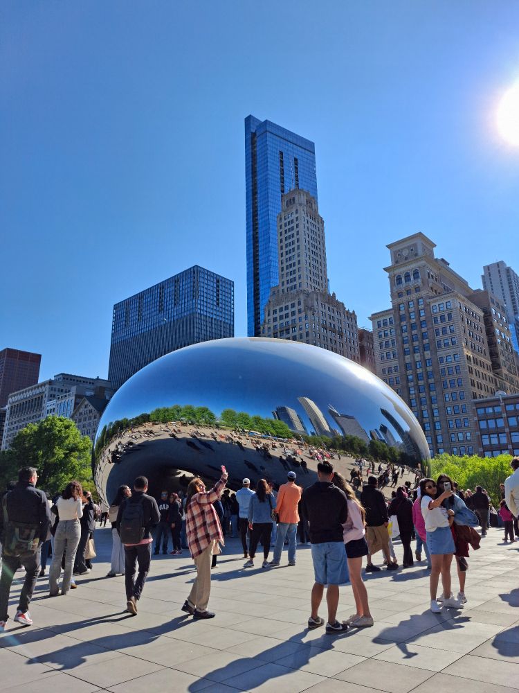 A large mirrored bean-shaped sculpture surrounded by people, green trees, and skyscrapers under a blue sky.
