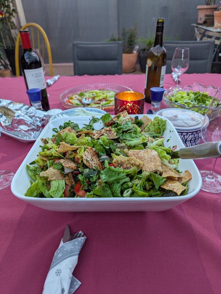 Fattoush salad in a large bowl, placed on a table set with a selection of salads, pebre and wine.