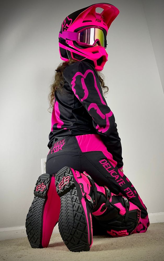 The racer pup kneeling from behind in his brilliant full pink-black 180 Fox Racing kit (pants are a knock off Delicate Fox, boots are comp 5 size 11, V1 Helmet. He playfully looks back at the camera on his right. His brown hair flows out of his helmet. Not shown but along his frontside he’s also wearing his pup pride bandana from Packappeal Apparel on Etsy and blue-black Mr S Leather collar.
