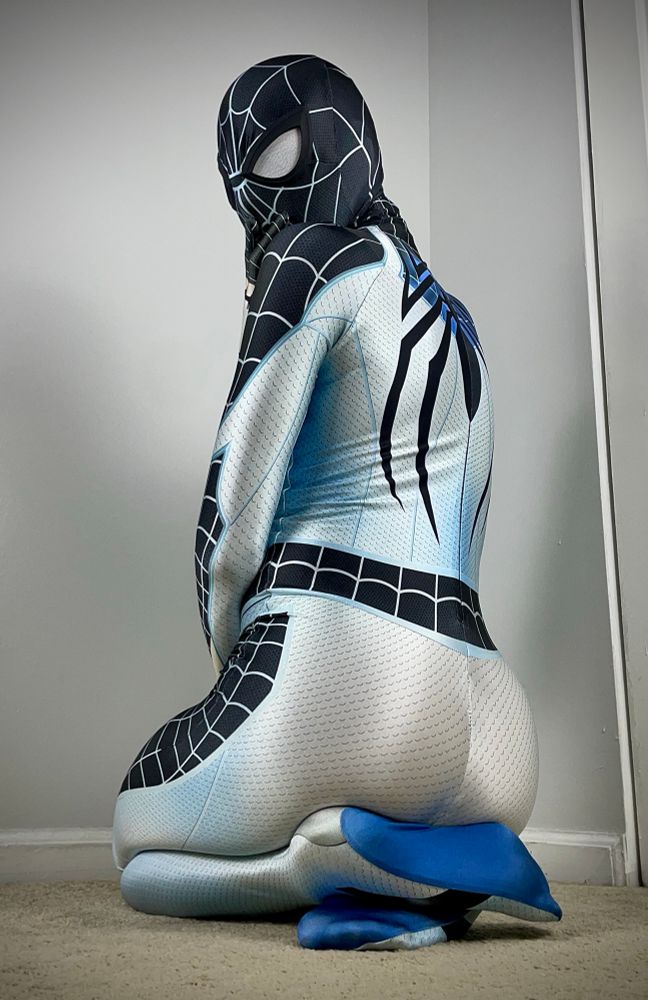 Spidey kneeled on his feeties with a view from his backside.He playfully looks back at the camera on his left side with his right hand covering his mouth. The suit features a slick pattern of light teal to blue contrasted with a black spider web pattern across his legs, arms, and mask. On his back (partially) is his blue spider emblem that also sits across his torso (out of view). (The suit is the Negative Spidey suit made from Zentai Hero).