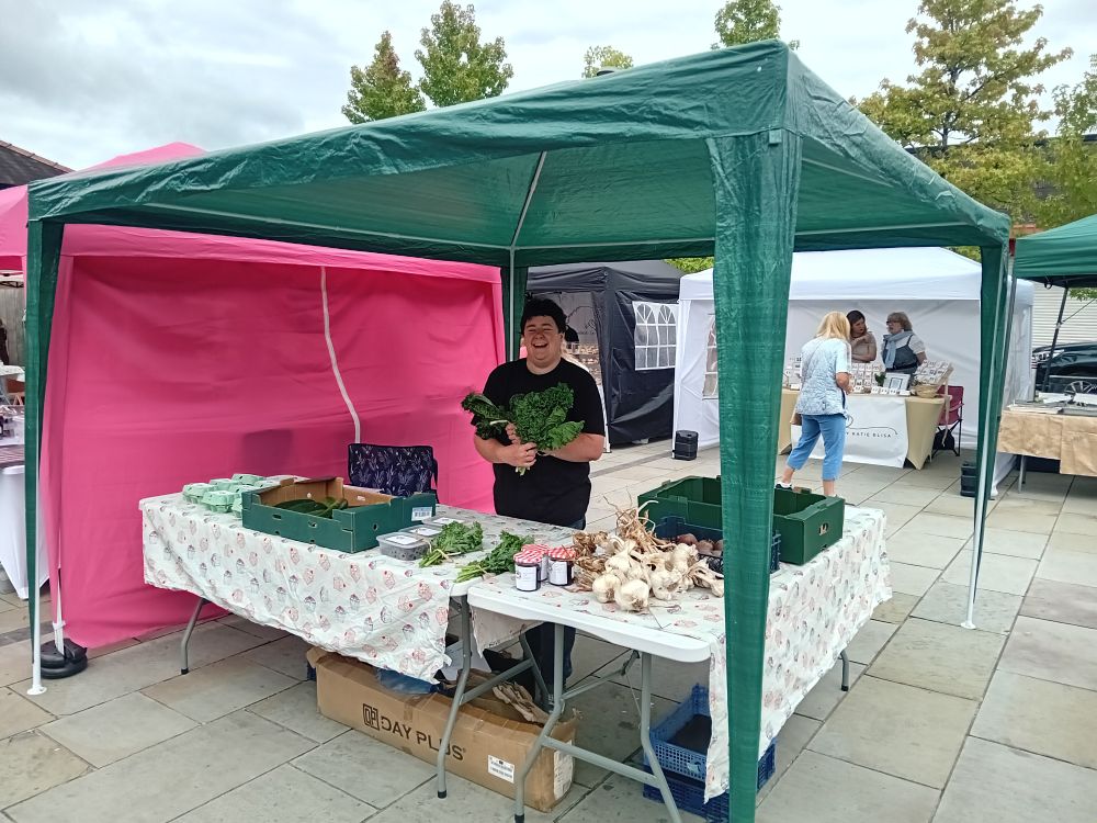 Me, white woman with dark hair wearing a black t shirt, standing under a green gazebo and behind a table covered in s tablecloth with cupcakes on it, holding two bunches of kale and looking jolly