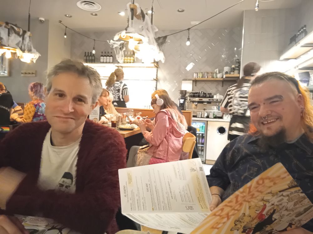 Chris - white man, red cardie, white t shirt and grey hair , is smiling and David, black top, balding, kind of orange hair, multiple facial piercings, is looking at a menu and laughing.