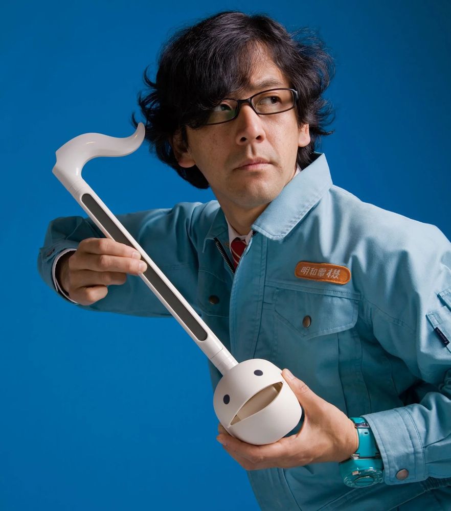 Image of the founder holding an atomotone, a digital synth type instrument. A simple picture with a plain blue background and the founder wearing a blue overall holding the instrument.