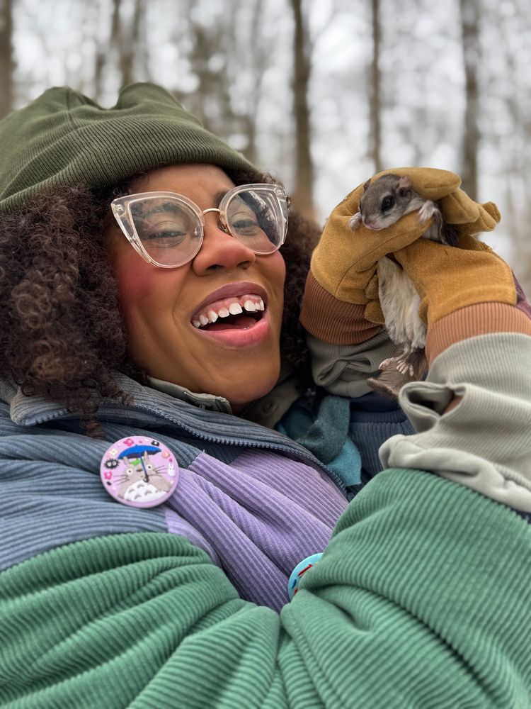 Alexis, a black woman with curly brown hair, wearing a beanie and corduroy jacket holds a southern flying squirrel 