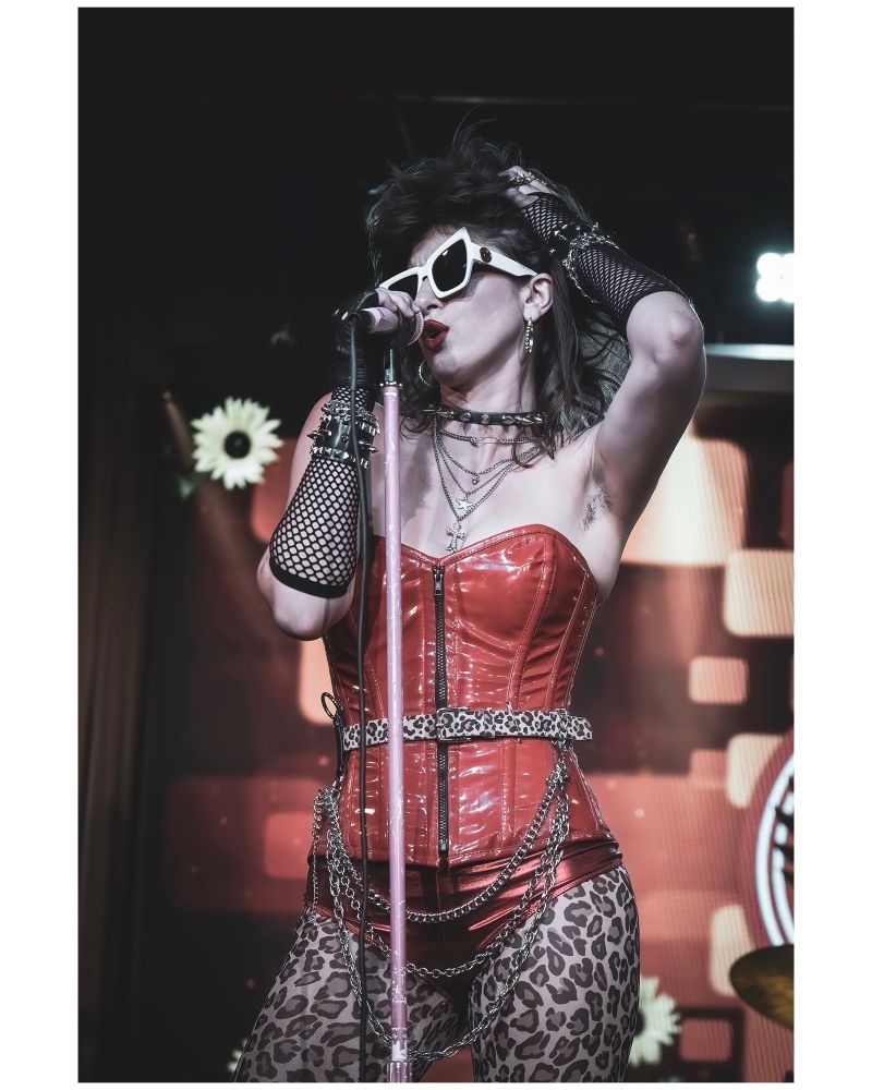 A color photo of lead singer Mikky Rib of Mikky & The DOOM wearing a red vinyl corset and asymmetrical white-framed sunglasses as she sings into a pink microphone on a pink stand.